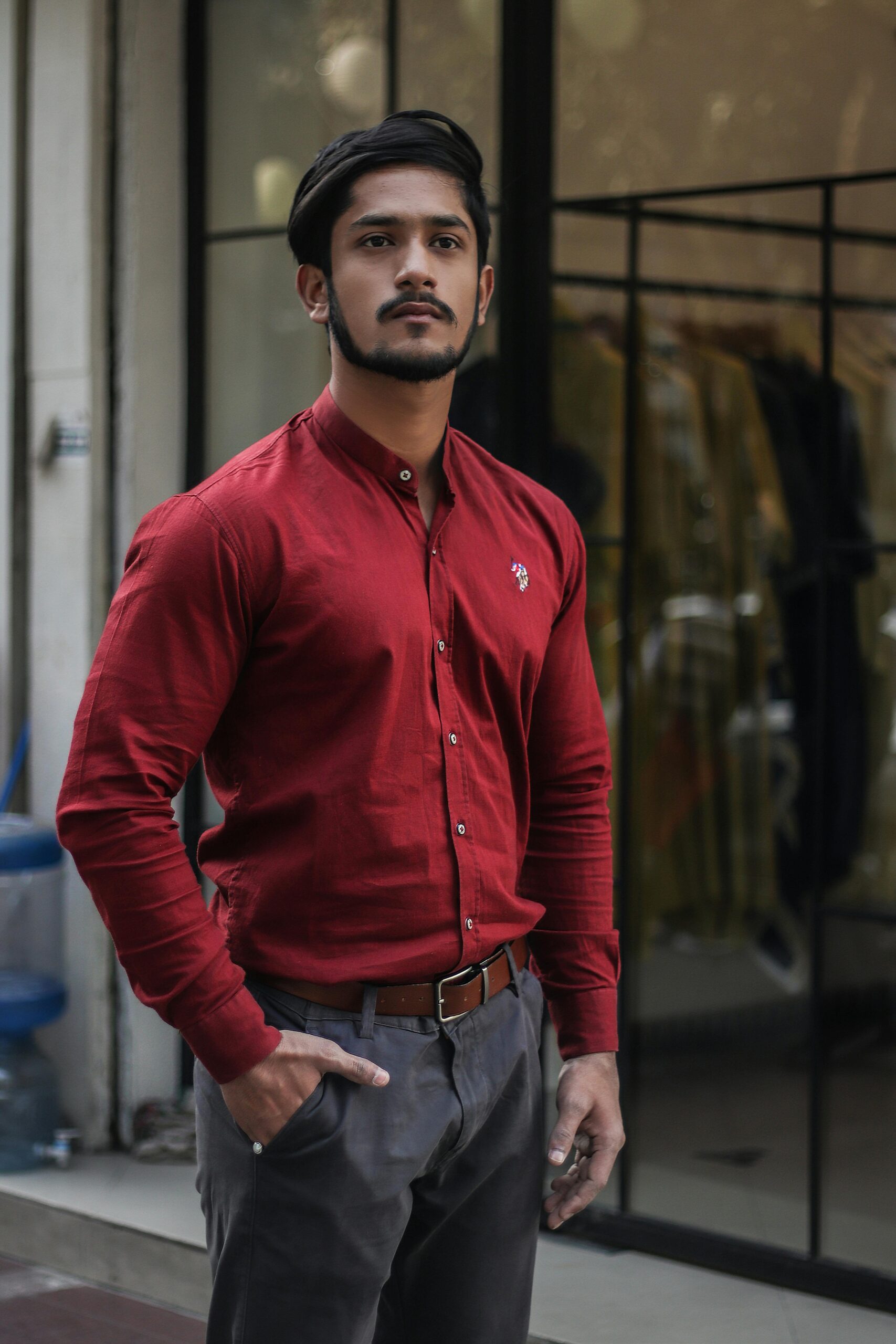 A stylish man poses confidently in a red shirt and gray trousers outside a clothing store.